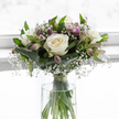 A calm, contemplative floral arrangement of white roses, gypsophila, and lavender alstroemeria is shown standing in a clear cylindrical glass vase, bathed in soft, even light that feels like a quiet morning in North Watford. The roses are generous and full, each bloom open to reveal layers of velvety petals that shift from a creamy ivory heart to clean white outer edges. Around them, delicate sprigs of baby's breath create a fine, cloud-like veil, so that the larger flowers appear to float in a gentle haze. Slender alstroemeria buds and partially opened blossoms introduce a muted wash of colour, with tones of lilac, soft lavender, and pale green, some petals subtly lined with fine, natural striping. Broad, deep green leaves are neatly folded and wrapped at the base, forming a protective cradle that makes the bouquet feel both grounded and lovingly held. Lighter foliage threads through the upper part of the design, bringing a sense of movement and life without breaking the calm. The clear vase, filled with fresh water, reveals the bundled green stems secured together with a small green tie, an unobtrusive but telling sign of skilled craftsmanship. The background is bright yet unobtrusive, allowing the bouquet's gentle lines and soothing colours to dominate the scene. This image captures a design that would be chosen to send comfort and respect-perhaps delivered to a family home off St Albans Road or to a small service nearby-offering a quiet message of support when someone in the North Watford community is going through loss.