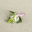 Displayed against a gently woven, light oatmeal linen background, this elegant boutonniere has the serene, polished look of flowers prepared for a meaningful celebration in North Watford. At its centre, a white Alstroemeria bloom opens generously, each petal soft and velvety, with fine reddish-brown stripes and tiny freckles running along the inner petals, drawing the eye inward as if inviting a closer look. Surrounding the main flower, a handful of smaller white buds and partially open blossoms suggest campanula or lily-of-the-valley, giving the piece a sense of purity and unfolding promise. Sprays of delicate white baby's breath weave between the blooms, their tiny stars creating a gentle, cloud-like halo that softens the design. On the left, a striking, bright green cone of tightly gathered Ornithogalum buds introduces an unexpected architectural element, its vivid colour contrasting beautifully with the creamy whites. The entire cluster is supported by broad, dark green leaves that fan out behind, grounding the boutonniere with lush, natural texture, while slender strands of grass-like foliage arc and curl around the edges, suggesting a breeze across Cassiobury Park or a walk toward a local church. At the base, the stems are tightly wrapped in a glossy pink satin ribbon, finished with a neat little bow that hints at romance, gratitude, or celebration. The lighting is soft and even, revealing every vein and contour without harshness, and the overall mood is one of calm joy and careful craftsmanship, as if created by a florist who understands both the emotion and the detail that a wedding or prom in North Watford deserves.