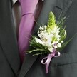 A close-up, high-angle photograph captures the upper torso of a man dressed for a formal event in North Watford, his charcoal grey suit jacket sharply tailored and perfectly framed in the shot. The fabric appears smooth and slightly matte, with the clean line of the lapel guiding the eye towards a carefully knotted lavender tie set against a pale dress shirt with a faint grid pattern. Pinned to the left lapel is a meticulously crafted wedding buttonhole, the true focal point of the image. At its crown rises a pale green, spire-like bloom of tightly closed buds, adding height and a sense of quiet anticipation, as if poised to open later in the day. Nestled below, several white Alstroemeria blossoms unfold, their petals marked with fine maroon and plum streaks that look like hand-drawn strokes, with tiny golden stamens glinting in the light. Wisps of white gypsophila, or baby's breath, hover around the main flowers, creating a soft, cloudlike halo that softens the strong lines of the suit. Slender, grass-like leaves curve outward in graceful arcs, while darker, broader foliage anchors the arrangement and deepens the contrast against the dark lapel. At the base, a satin lilac ribbon is tied into a neat bow, its silky sheen echoing the cool tone of the tie and adding a gentle romantic flourish. The overall mood is one of poised celebration, the kind of refined boutonniere a groom might wear to a wedding at a local North Watford church or registry office, expertly arranged to look timeless in photographs and quietly special in person.