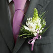 In this detailed close-up, the photograph lingers on the left lapel of a dark charcoal suit, capturing the quiet drama of a single boutonniere prepared for a ceremony somewhere in North Watford, perhaps not far from Garston or the quiet streets around Watford Junction. The jacket fabric appears rich and finely woven, its lapel lying flat in a clean line over a pale shirt patterned with a barely-there dark check that adds subtle depth without stealing attention. Centre stage is a smooth lavender necktie, knotted traditionally and falling in a straight, unbroken line, its satiny sheen catching light like evening sun glancing across the Grand Union Canal. Pinned just beside the tie knot, the boutonniere brings the whole look to life. Several white alstroemeria blossoms unfurl, each petal showing slender reddish-brown brushstrokes and soft yellow throats, their stamens clearly defined. Rising from the heart of the arrangement, a fresh green, cone-shaped bud introduces a slightly architectural feel, while tiny clusters of white gypsophila nestle between the larger blooms like flecks of foam. Around them, a mix of slender, spiky greenery and broader, glossy leaves frames the flowers, offering layers of texture that a hand could trace from smooth to feather-light. At the base, a lilac satin ribbon is tied into a neat, symmetrical bow, its colour perfectly echoing the lavender tie so the whole palette feels coordinated yet gentle. The mood is one of poised celebration-a floral detail chosen with care, ready to walk down an aisle in North Watford or share the spotlight at a prom or black-tie event, where every small element helps tell the story of the day.