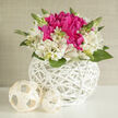 This elegant floral design is captured against a bright, uncluttered background, giving each blossom space to breathe while still feeling intimately gathered, much like a quiet celebration in a North Watford townhouse. At its core, a rounded cluster of fuchsia peonies, or peony-style blooms, swirls with densely layered petals, the colour deepening toward the centre into velvety magenta shadows. Around this vivid core, creamy white alstroemeria arc outward in a loose ring, their trumpet-shaped petals kissed with fine magenta streaks and soft yellow throats, creating a halo of lightness and air. Slender green stems and buds rise between the flowers, some just beginning to open, suggesting new beginnings and the gentle unfolding of a special day. The arrangement is nestled in a round, snow-white wicker basket, woven in sleek, overlapping strands that form a delicate lattice, almost like a sculpted piece of modern art. The wicker's cool tone and tactile pattern contrast beautifully with the softness of the petals, giving the eye a resting place and anchoring the design. Two softly woven cream spheres sit close by, echoing the basket's texture and adding an understated decorative accent, as if styled on a coffee table in a light-filled living room near St Albans Road. The lighting is calm and diffused, creating subtle shadows that enhance each petal's curve and every vein in the leaves, evoking a tranquil yet uplifting mood. This arrangement feels ideal for bridal showers, intimate wedding collections, or to honour milestones with quiet sophistication. As a local florist, I can almost imagine this bouquet arriving with same-day delivery in North Watford, bringing a whisper of fresh fragrance and thoughtful beauty into the room.