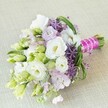 A graceful, hand-tied bouquet rests atop a subtly textured beige background, capturing the spirit of North Watford's springtime celebrations. The arrangement, lovingly crafted by a local florist, showcases ruffled creamy white Lisianthus blossoms as the focal point, surrounded by slender, arching stems of bright green bear grass and feathery foliage, evocative of the tranquil corners of Callowland Park. Intermixed are gentle clusters of pastel lavender-pink stock flowers and vivid deep purple alliums-each star-shaped floret sparkling in the soft daylight. Tightly bound stems are wrapped in a bold fuchsia satin ribbon and adorned with a strand of pearlescent beads, adding both contemporary flair and timeless elegance. Sprigs of white and lime-green Lisianthus buds drift between the main blooms, suggesting natural growth and fresh beginnings. The bouquet feels vibrant yet refined, bringing together the romance and optimism of a North Watford wedding morning. It's an arrangement designed for joyful occasions, blending classic beauty with modern touches, and ideal for those celebrating life and love within this welcoming Hertfordshire community.
