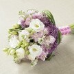 A beautifully composed, hand-tied bouquet rests on a neutral, softly textured background, its palette a harmonious blend of soft lavender, crisp white, and fresh green reminiscent of North Watford's tranquil green spaces. Prominent among the flowers are several full, ruffled white Lisianthus, their luxuriant petals softly encircling creamy yellow centres. Interlaced within are clusters of delicate lavender Stock, adding an airy froth of colour and gentle fragrance, expertly complemented by bright green, arching foliage and the occasional unopened Lisianthus bud. The arrangement's round, slightly elongated form provides visual depth and classic sophistication, perfect for weddings or elegant events around the Watford Junction area. Binding the stems is a pastel-shaded pink ribbon, subtly striped in white and enhanced by a tactile strand of pearls-a finishing detail echoing the elegant style favoured by North Watford's couples. Bathed in even, natural light, every textured petal and leaf is revealed in exquisite detail, making this bouquet an inspired focal point for bridal celebrations, anniversary dinners, or anniversary gifts delivered fresh to homes across the local community.