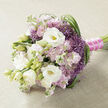A refined wedding bouquet is displayed lengthways on a pale beige, slightly textured fabric that resembles soft linen, providing a calm, neutral canvas much like the quiet pavements around St Albans Road in North Watford. The arrangement blends white, blush pink, and lilac-purple tones into a harmonious, romantic palette. At its centre, generous white lisianthus blossoms unfold in layers, their petals silky and rounded, with a hint of yellow-green peeking from each throat. Some lisianthus are fully open, while others remain in smooth, elongated buds, adding depth and the promise of blooms yet to come. Around them, clusters of pastel stock flowers in soft pink and light mauve create a pillowy effect, each stem bursting with small, ruffled blooms that bring a sense of gentle abundance and a faint, sweet fragrance. Interlaced throughout are starry purple filler flowers, reminiscent of ornamental allium, their spiky, spherical heads introducing texture and a lovely contrast of shape and colour. Slim blades of deep green foliage weave through the bouquet, looping and arching gracefully, especially along the right-hand side, guiding the eye and giving the whole design a natural, flowing rhythm that feels almost like a walk through a spring garden near North Watford's park edges. The gathered stems are neatly wrapped in a vivid fuchsia satin ribbon, which stands out against the softer tones above. A delicate strand of tiny white pearls is pinned along the ribbon, catching the light with subtle highlights and suggesting bridal jewellery. The bouquet radiates poise, romance, and modern elegance, ideal for a North Watford bride stepping into her ceremony, a special anniversary surprise, or a refined celebration where understated luxury is cherished.