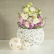 A bright, contemporary floral arrangement, crafted with the expertise of a North Watford florist, graces a spotless white tabletop, bringing warmth and elegance to its setting. Central to the display are abundant creamy white and pale yellow eustoma, their ruffled petals layered like the blooms found in gardens near North Watford Recreation Ground. Sprays of soft pink stock flowers rise gracefully, infusing the bouquet with gentle colour and a sense of height. Underneath, clusters of radiant purple fillers-perhaps aster or small allium-provide textural richness reminiscent of countryside hedgerows. A ring of deep green foliage outlines the base, its lushness highlighting the pastel blooms above. The flowers are presented in a striking white spherical vase, its open-weave construction from painted rattan or intertwined branches lending a touch of artistic flair, reminiscent of North Watford's creativity and handmade traditions. To the left, two decorative woven orbs crafted from lighter natural wood sit side by side, their airy basket-like patterns reflecting the vase's structure, adding cohesion and tactile interest. The neutral greige wall behind the arrangement, with its understated vertical texture, enhances the tranquil, sophisticated ambiance. Bathed in soft, natural light, this arrangement conveys a serene yet inviting mood-ideal for spring gatherings, wedding centrepieces, or as a stylish statement for any special event throughout North Watford. Each detail reflects the care and artistry found within the local floral community.