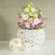 On a smooth white tabletop, a graceful floral display becomes the quiet focal point, as if waiting to greet someone stepping in from the streets of North Watford after a long day. At the centre, generous lisianthus blooms in soft white and warm cream-yellow unfold in multiple silky layers, their petals curling gently outward with a romantic ease. Around them, clouds of blush pink stock cluster closely, each spike made up of many petite blossoms that lend a sense of softness and gentle fragrance. Interspersed are smaller, spherical purple allium heads, their tiny starry flowers creating a subtle, spiky texture that balances the otherwise velvety feel. Lush green foliage is woven throughout the arrangement, with a deliberate band of darker leaves encircling the base like a natural ribbon, visually gathering the bouquet into a rounded, abundant form. The flowers are cradled in a distinctive white spherical vase, crafted from slender branches or twigs arranged into a light, airy lattice, allowing glimpses of stems and shadows inside the orb. Its bright white tone contrasts sharply with the pastel flowers and green foliage, giving a fresh, gallery-like impression that would suit a modern flat near North Watford's parklands or a minimalist office space. To the left of the vase, two decorative orbs made from pale, interlaced wooden strips echo the woven structure, one larger and one smaller, adding rhythm and a touch of rustic warmth. Behind, a softly blurred wall in a muted beige or greige texture acts as a calm backdrop. The overall mood is tranquil and contemporary, a thoughtful composition perfect for marking quiet celebrations, offering comfort, or styling an elegant interior.
