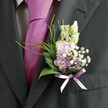 A close-up photograph focuses on the upper torso of a man dressed for a formal occasion in North Watford, showing only his charcoal suit jacket, patterned shirt, and carefully chosen buttonhole. The suit is a deep charcoal grey, almost black, with a soft, subtly textured fabric that catches the natural light along the lapel and sleeve. Beneath it, a crisp, pale dress shirt with a fine grid check peeks through, offering a gentle contrast in both colour and pattern. At the centre of the image, a bold magenta silk tie, neatly knotted, glows with a lustrous sheen, its smooth surface reflecting the soft daylight in a way that suggests a spring or early summer wedding morning. Pinned precisely to the left lapel is an intricate boutonnière, the true focal point of the scene. Slender, creamy white buds-reminiscent of snapdragons-rise at the top, leading the eye down into small clusters of blush pink and lilac-lavender blooms that could be sweet peas or delicate stock. Tiny white clouds of gypsophila nestle between them, adding a misty, romantic lightness, while a few deeper purple florets introduce depth and nuance. At the base, glossy dark green leaves create a solid foundation, offset by fine, feathery fronds of brighter greenery that arc outward like soft, airy brushstrokes. A narrow violet-lavender satin ribbon holds the stems in a neat bow, echoing the cooler purple tones in the flowers and giving the design a polished finish. The image conveys a sense of quiet confidence and joy, the kind of refined detail you might see on a groom stepping out from a townhouse near Cassiobury Park or a smart venue off St Albans Road, ready for a heartfelt celebration.