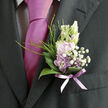 Pinned neatly to the lapel of a dark, tailored suit, this delicate boutonnière offers a soft counterpart to the crisp lines of formalwear, much like a gentle breeze drifting across North Watford on a clear spring afternoon. At its centre, pale lavender blossoms open in layered petals, their colour a quiet whisper of romance that harmonises beautifully with the deeper tones of the nearby tie. Surrounding these main flowers, tufts of tiny white filler blooms-reminiscent of gypsophila or baby's breath-form a light, cloudlike frame, adding texture and a sense of airy movement. Glossy green leaves curl around the base, their surfaces smooth and slightly reflective, creating a natural cradle that keeps the arrangement tidy yet organic. Slender blades of fresh green grass lift upward from behind the blossoms, giving the composition a lively vertical rhythm, as though the flowers were still reaching for the sun above the gardens off St Albans Road or along the paths near Knutsford Playing Fields. The stems are bound together with a sheer lavender ribbon, its soft shimmer catching any available light and subtly echoing the overall colour palette. Texture plays a key role: the ribbon's satin touch contrasts with the cool wool of the suit and the delicate, almost papery quality of the petals, suggesting both refinement and sensitivity. This small floral piece feels like a thoughtfully chosen finishing touch for a wedding, prom, or elegant celebration, perhaps delivered locally by Flower Delivery North Watford to a venue tucked near Garston or around the busy heart of the town. The boutonnière conveys grace, composure, and personal sentiment in a compact, beautifully balanced design.