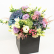 A vibrant, opulent floral bouquet-brimming with colours and textures-stands in sharp relief against a crisp white background, elegantly arranged in a sleek, matte black square box. This garden-style masterpiece, crafted by florists in North Watford, features abundant clusters of deep blue and periwinkle hydrangeas, their pillowy petals forming a luminous base. Intertwined are soft lavender and blush pink roses, each bloom delicately unfurling to convey warmth and romantic joy, while creamy white lisianthus and buttery yellow eustoma trumpet upward, adding volume and a subtle glow, much like sunshine gracing local parks. Sprays of bright green 'Green Trick' dianthus nestle among the blossoms, injecting a striking modern accent and tactile contrast. Wispy plumes of light pink astilbe offer playful movement and soft nostalgia, reminiscent of home gardens in North Watford. The arrangement's lush, varied greenery-from glossy deep leaves to zesty lime and newly sprouting buds-fills each corner, bringing fresh, uplifting vibrancy. Balanced expertly, the mix of cool purples, blues, airy creams, and bright greens is framed by the understated elegance of the minimalist black container, allowing the flowers' richness to command attention. Bathed in natural, even light, every colour radiates its beauty, making this design perfect for thoughtful gifts, elegant home décor, or articles highlighting luxury flower delivery and unique gift ideas across North Watford.