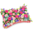 A pillow-shaped sympathy tribute captured from a slightly elevated, oblique angle against a clean white backdrop, this densely packed floral cushion reads both intimate and celebratory. The surface is a plush tapestry of hot pink roses-some tightly furled, others gently unfurling to reveal velvety centers-set beside clusters of soft, shell-pink carnations that lend a ruffled, tactile softness. Bright lemon-yellow ranunculus punctuate the composition with sunny, buttercup-like heads, while deep purple lisianthus (eustoma) add bell-shaped contrast and a graceful depth. Scattered through the blooms are silver-blue eryngium thistles, their architectural, spiky forms introducing a cool, wild edge against the otherwise rounded petals. Rounded eucalyptus leaves with their silvery-green sheen and assorted darker greens weave through the arrangement, giving it dimensional shadow and a faint, calming scent of fresh foliage. Encircling the pillow is a rich red pleated satin ribbon, folded into a ruffled border that both defines the rectangular cushion silhouette and adds an elegant textural frame. The lighting is soft and natural, allowing petal surfaces to catch and reflect a subtle sheen while keeping the overall mood comforting rather than austere. As a floral pillow intended for funeral or memorial presentation, the palette-fuchsia, lavender, lemon, and blush-speaks of affection and remembered joy rather than stark mourning, offering warmth and consolation. A local florist's hand is evident in the careful balance of color, the density of placement that preserves the cushion's form, and the thoughtful use of contrasting textures to convey a message of love and gentle remembrance in the North Watford community.