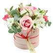 A luxurious round hatbox bouquet in soft pastel tones is presented against a clean white background, capturing the look of a premium floral gift ready to be delivered across North Watford. The box is cylindrical and finished in matte blush pink, giving it a gentle, modern look. Around its middle runs a slim coral-red satin ribbon, carefully tied into a neat little bow at the front, from which a cream-coloured gift tag with elegant green writing and a small web address lightly dangles, hinting at a personalised message from the sender. Overflowing from the top of the box is a lush, domed arrangement of fresh flowers in shades of pink, white, and green. At the heart of the bouquet sits a striking, fully opened rose with pale outer petals and a more intense pink swirl at its centre, creating a focal point of velvet-like texture. Surrounding it are clusters of white chrysanthemum-style blooms with bright green centres, their tidy, daisy-like faces adding both structure and brightness, reminiscent of neat front gardens off St Albans Road. Delicate white alstroemeria with slender petals and fine speckling thread gracefully between the larger flowers, while smaller, deep pink blossoms-possibly spray roses or lisianthus-are tucked in for flashes of rich colour and romance. The composition is softened and framed by generous greenery: rounded, silver-green eucalyptus leaves lend a cool, contemporary feel and suggest a fresh, clean fragrance, while darker green foliage and tiny green buds offer additional layers and natural movement. The arrangement feels polished yet inviting, ideal for marking birthdays, anniversaries, Mother's Day, or simply sending a heartfelt gesture to someone special in North Watford.