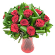A luxurious, tightly rounded bouquet of roughly a dozen deep crimson roses fills the frame, each bloom open to reveal velvety, spiraled petals that catch soft, even light and glow almost wine-dark. The roses are tucked into a generous skirt of glossy, dark green foliage - flat Ruscus- or Salal-like leaves forming a dense, verdant backdrop while broader, Aspidistra-style leaves are sculpted into elegant curls and spirals that loop around the heads like architectural ribbons. Those folded green shapes add a contemporary, structured rhythm against the classic rose heads, creating movement and balance from stem to tip. The whole composition sits in a tall, translucent ruby-red glass vase whose subtle ribbed or twisted surface throws vertical highlights and echoes the roses' passionate tones; the vase narrows slightly at the waist and flares gently at the mouth, grounding the arrangement with quiet poise. Photographed against a pristine white background, the intense reds and rich greens are isolated and amplified, allowing the bouquet's textures - the matte softness of petals, the glossy sheen of leaves, the glinting glass - to be read clearly at a glance. There is the faint suggestion of a classic rose scent, warm and reassuring, a scent meant for anniversaries, Valentine's Day, or any moment when heartfelt feeling must be conveyed. As a local florist in North Watford I picture this arrangement arriving at a family table near Cassiobury Park or sent as a romantic surprise to a home off Watford High Street; its timeless elegance and modern leaf sculpting suit both intimate gifts and refined centrepiece duties.