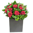 A contemporary composition of deep, velvety red roses clustered at the heart of a low, matte black cube; each bloom fans open in generous spirals, the petals soft and satiny under bright, even studio light. Surrounding the roses, broad glossy green leaves have been carefully curled and overlapped, creating a rhythmic movement that frames the crimson centers and lends sculptural depth to the design. The square vase's textured matte surface provides a steady, modern counterpoint to the lushness above, its geometry anchoring the bouquet like a sculpture placed on a city mantel. Lighting catches the sheen on every leaf and the subtle highlights on the rose petals, giving the arrangement a fresh, just-unwrapped look that suggests morning light on a clean windowsill. If you lean in, the scent reads clearly as classic rose-softly sweet and heady-lifted by crisp green notes from the foliage, a combination that feels both intimate and invigorating. As a local florist I picture this piece on a dining table for an anniversary, beside a bedside for a loving surprise, or brightening an urban office near Cassiobury Park; it radiates romance and quiet confidence without fuss. The craftsmanship is deliberate: each leaf loop, each bloom placement creates balance and movement, transforming simple elements into a bold but refined statement that fits modern interiors across North Watford.