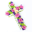 A beautifully composed floral cross stretches diagonally across a brilliant white background, its careful construction and vibrant colours combining to create a meaningful funeral or memorial tribute for a North Watford service. The outline of the cross is crisply defined by a thick edging of tightly packed white spray chrysanthemums, each tiny flower contributing to a soft, cloud-like frame that gently cradles the design. Just outside this white halo, a slim band of light, fresh green-perhaps a folded satin ribbon or neatly wrapped foliage-peeks around the perimeter, adding a subtle, tailored finish and a suggestion of new life. Within this frame, the interior of the cross is luxuriously filled with mixed blooms. Large roses in bright fuchsia pink and soft creamy yellow are fully opened, their spiral petals forming lush focal points along the vertical centre and outward across the arms. Dotted among these bold roses are clusters of lime-green button chrysanthemums, their rounded, pom-pom heads offering a lively texture and a modern burst of colour. Interwoven throughout are smaller flowers in deep purple and violet tones, such as statice or lisianthus, which slip between the larger blooms and lend depth and contrast. The result is a carefully balanced palette of pink, cream, green, and purple that feels both comforting and hopeful. Strong, even lighting highlights every petal edge and the gentle sheen of the green border, while the white backdrop keeps the focus firmly on the cross. This arrangement evokes love, remembrance, and quiet faith, perfect for honouring a loved one at a local church or crematorium in the North Watford community.