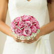 In this tender bridal scene, a rounded bouquet of blush pink roses and airy white gypsophila rests securely in the bride's hands, becoming a graceful focal point against her lace wedding dress somewhere in North Watford. Each rose is at its prime, petals unfurling in layered spirals from deep blush at the centre to soft shell-pink near the edges, creating a gentle ombré effect that looks almost painted. Wisps of baby's breath weave between the roses, their tiny white blossoms like soft snowflakes, blurring the boundary between flowers and fabric. The bouquet is compact yet full, perfectly symmetrical, giving a sense of calm order and quiet confidence as the bride prepares to walk down the aisle. Her gown, rich with ivory lace and delicate patterning, forms a textured backdrop that reflects the natural daylight, perhaps streaming in from a venue near Garston or along St Albans Road. The interplay of light and shadow accentuates every curve of the petals and every tiny bloom of gypsophila, creating a gentle, dreamlike glow. The scent is subtle and romantic: the fresh sweetness of garden roses, with just a hint of green from the stems and foliage hidden beneath the blooms. This is the kind of bouquet a local florist designs with real care, imagining the bride stepping out of a car near a North Watford church, the bouquet steady in her hands as her heart flutters with anticipation. Everything about the composition-its softness, balance, and pastel palette-speaks of enduring affection, new beginnings, and a love story unfolding one step at a time.