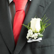 In the softened daylight that slips into a wedding reception or formal photo, an ivory rose boutonnière sits with confident restraint against a smart charcoal jacket. The rose's velvety petals open in layered spirals, their creamy tones catching subtle highlights that suggest coolness and a tactile softness beneath the fingertips. Behind the bloom, lacy sprays of baby's breath scatter like tiny stars, their cloud-like texture balancing the rose's solidity with a whisper of innocence. A checked shirt peeks beneath the lapel and a bold red silk tie adds a romantic punctuation, the contrast between textile and flower lending the ensemble a timeless, storybook quality. Fine slivers of fern and twisted leaves arch around the stem, introducing movement and a painterly depth while their glossy surfaces reflect narrow shafts of light. You can almost sense the morning dew on the petals and the faint, familiar scent of fresh rose that rises when someone leans in to offer congratulations. This boutonnière feels handcrafted for a groom or honoured guest moving through ceremonies at Vicarage Road or meeting friends for photos by Watford Junction - an understated but meaningful accent that complements both formal tailoring and the warm rhythms of North Watford celebrations.