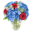A striking hand-tied bouquet sits in a simple clear glass vase, photographed against a clean white backdrop so every texture and colour reads clearly. The composition is anchored by generous clusters of sky-blue hydrangeas whose many tiny petals form soft, cloud-like mounds that catch bright, natural light; their cool, watery blue suggests a summer sky just after rain. Threaded through that plush blue are three velvety red roses, their petals tightly spiralled and richly saturated, creating a dramatic, romantic punctuation that draws the eye inward. Scattered like tiny stars throughout the mass of blooms are delicate sprays of white baby's breath, their feathery silhouettes adding an airy, ethereal softness that lightens the overall shape. Muted green eucalyptus leaves with rounded, matte surfaces weave between the flowers, offering a subtle herbal scent and a calm, silvery-green counterpoint to the saturated blues and reds. The vase's gently rounded base and tapered mouth reveal the fresh green stems submerged in clear water, a visual cue to the bouquet's freshness and the careful mechanics of floristry. The entire arrangement is brightly lit from above and slightly to the side, which highlights petal texture, the velvet of the roses, and the soft sheen on the hydrangea leaves. As a local florist would describe it to a customer, this piece feels both celebratory and intimate-perfect as a centerpiece for a birthday table, an anniversary surprise, or a patriotic display for a community event in North Watford near Cassiobury Park-radiating confident colour while remaining gracefully composed.