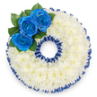 A bespoke memorial wreath formed into a perfect circular '0' shape, photographed from above against a clean white backdrop, radiates quiet dignity and calm. The entire number is built from a dense, pillowy carpet of small white spray chrysanthemum florets - hundreds of tiny, velvety petals packed so closely they read as a soft, luminous surface that suggests peace and gentle consolation. Around both the outer and inner edges a pleated royal blue satin ribbon has been carefully machine-stitched and hand-ruffled to follow the contours; its scalloped trim mirrors the wreath's roundness and catches the light with a subtle sheen that echoes the blue of the roses. Anchoring the upper-left curve is a focused cluster of three deep royal blue roses, their buds opening outward in layered spirals of velvety petals; these vivid blooms are set against a few glossy, dark green leaves that add a natural depth and a cool contrast to the white cushion beneath. The blue used here is deliberate - a tranquil, almost oceanic tone chosen to convey serenity, trust, or to reflect a cherished colour associated with the person being remembered. Soft studio lighting throws gentle shadows into the pleats and petals, revealing texture without harshness and keeping the arrangement contemplative rather than theatrical. As a local florist's tribute, this numbered wreath is often requested for milestone memorials and funeral services across North Watford - from homes near Cassiobury Park to community halls along St Albans Road - offering a personalised, respectful emblem of remembrance.