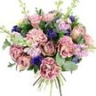 A richly detailed hand-tied bouquet is shown against a bright white background, every stem and petal arranged with the care of an experienced North Watford florist. The eye is first drawn to the generous garden roses at the centre, their full blooms opening in soft spirals of blush pink, dusty mauve, and antique rose, evoking a gentle, timeless romance. Encircling these are layers of ruffled carnations, each petal edged with frills that catch the light. Their colouring shifts from creamy, almost beige centres to soft blush and then to livelier fuchsia and plum-pink tones at the tips, giving the bouquet a sense of depth and movement. Between the warmer shades sit cool, jewel-like accents: closed, tulip-shaped buds in deep indigo and a single royal blue anemone with a dark, almost black centre, placed near the lower front like a focal gemstone. Slim spires of lilac and pale lavender stock rise delicately among the other stems, bringing a soft, floral scent and a touch of cottage-garden charm reminiscent of borders in nearby parks and green spaces around North Watford. These blooms rest in a bed of mixed greenery, including glossy dark leaves and the distinctive, rounded discs of silvery eucalyptus that arc around the bouquet, suggesting a refreshing, aromatic note. The neatly bound stems are visible at the base, ready to be placed in water or delivered as a thoughtful gesture across North Watford, whether to mark a birthday, send congratulations, or offer quiet comfort. The overall impression is one of calm elegance and tender emotion, captured through layered textures and harmonised colour.