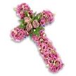 A thoughtfully crafted floral cross tribute is shown in full, resting visually against a bright white background that makes every colour and detail stand out. The cross itself is lush and three-dimensional, completely covered with flowers so that the underlying frame is almost entirely hidden. Soft pinks, dusty mauves, and delicate purples dominate the palette, creating an atmosphere of tenderness and calm. At the intersection of the cross, three large roses in a muted rose-lavender shade form a striking centrepiece, their softly spiralling petals giving the sense of a gentle embrace. Around them, airy fern fronds and compact green leaves spread outward, adding freshness and a touch of woodland character that feels familiar to anyone walking through North Watford's parks on a quiet afternoon. Flowing from this central cluster, the arms and length of the cross are densely packed with lisianthus blooms, their ruffled, trumpet-shaped petals overlapping in layers of blush pink, deeper rose, and lilac, some showing darker eyes at their centres. Nestled among these are small pink and purple filler flowers, creamy-white blossoms, and pale green buds that contribute fine detail and varied texture, like a choir of softer voices supporting the main theme. Along the outer outline and at a few recesses in the design, glimpses of deep purple ribbon or base material discreetly frame the flower mass, reinforcing the shape without intruding on its softness. The balance of colour, form, and foliage conveys a strong sense of peace and reverence. This is the kind of floral cross a North Watford family might choose to send as a final tribute, symbolising faith, love, and enduring remembrance during a funeral or memorial service.