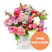 A carefully composed floral bouquet appears against a bright white background, allowing every colour and petal to stand out with clarity. At the centre are several striking roses in shades of pink, from deep fuchsia with darker edges to softer, more delicate blush tones that suggest tenderness and affection. These roses are woven together with generous carnation blooms in coral, salmon, and peach-pink, their layers of ruffled petals giving the bouquet a plush, almost cushion-like softness. Between the larger flowers, small daisy-like chrysanthemums in lavender and purple peek through, each highlighted by vivid green centres that add a fresh, zesty spark to the composition. Pockets of creamy white blooms are thoughtfully positioned to soften and balance the strong pinks and purples, keeping the overall look light and approachable. A rich mix of foliage frames the flowers: rounded eucalyptus leaves in silvery blue-green tones, deeper green sprigs, and touches of reddish-brown leaves create depth, dimension, and a subtle hint of autumn warmth. The flowers are arranged in a clear, square glass vase with slightly rounded corners, giving a modern yet timeless feel. Around the base of the vase, a white decorative band is wrapped neatly, printed with tiny red hearts and the affectionate phrase all my love, turning the container into part of the message. In the lower right of the image, an orange circular graphic clearly displays the words VASE INCLUDED in bold white capitals, signalling that the arrangement is delivered ready to place on a table or mantel. The bouquet feels like a thoughtful surprise you might send to a loved one in North Watford, perhaps to brighten a home near Garston or along Bushey Mill Lane, suitable for birthdays, romantic gestures, or heartfelt thank-yous.