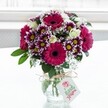 A vibrant mixed bouquet in a clear, textured glass vase sits centered against a softly glowing white background, as if placed on a bright North Watford windowsill on a calm morning. The arrangement is dominated by large gerbera daisies in deep fuchsia and magenta, their velvety petals circling dark, inky centers that immediately draw the eye. Around them, multi-petalled chrysanthemums in rich purple with white-tipped edges form a halo of colour; each daisy-like face has a sunny yellow heart that seems to sparkle under the natural light. Nestled between these bolder blooms are creamy white roses, some fully open with layered, satin-soft petals, others still in tight buds suggesting freshness and new beginnings. Fine sprays of white gypsophila, or baby's breath, are threaded delicately throughout the bouquet, creating a cloud-like softness and adding a romantic, airy texture. Deep green foliage frames the flowers and spills lightly over the rim of the vase, grounding the vivid palette with a natural, garden-fresh feel. The squat vase itself is cinched at the neck with a simple bow of rustic twine, from which hangs a small white rectangular gift tag featuring a pink floral emblem and hints of red script, suggesting a handwritten message for a birthday, thank you, or just-because surprise. The overall mood of the image is joyful and celebratory, like a thoughtfully chosen floral gift just delivered to a home off St Albans Road or near Cassiobury Park, ready to brighten a kitchen table. Subtle reflections in the glass hint at the stems gathered neatly inside, reinforcing the sense of professional care and attention that a local North Watford florist brings to each arrangement, while the bright, slightly blurred background keeps the focus firmly on the flowers' vivid colours and lush textures.