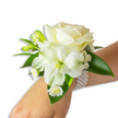 A delicate, handcrafted white wrist corsage rests on a light-skinned wrist, photographed against a clean white background so every texture reads clearly. At the heart of the piece sits a creamy ivory rose, its velvety petals unfurling in soft concentric layers that catch the light with a subtle, pearlescent sheen. Just below and a touch left, a trumpet-shaped white alstroemeria (Peruvian lily) expands outward, its slender petals revealing a pale green-yellow throat that gives the arrangement gentle depth. Tiny, airy sprigs of Gypsophila (baby's breath) halo the main blooms, their cloud-like clusters creating a whisper-soft edge, while small daisy-like white florets with golden-yellow centres tuck in near the base to add a cheerful, intimate note. Several fresh green buds, still tightly rolled, promise movement and a future bloom, and broad, glossy deep-green leaves with a smooth waxy finish frame the composition and provide a rich contrast to the pristine whites. The whole floral cluster is elegantly anchored by a multi-strand pearl wristlet: uniformly round, luminous faux pearls wrap the wrist, offering a cool, tactile counterpoint to the petals. If you imagine raising that wrist to greet someone at a wedding or prom, the corsage emits a faint, powdery floral scent-clean, soft, and reassuring-soothing in the way crafted florals can be. As a local florist's piece conceived for quiet sophistication, the design balances classic formality with a just-picked freshness, ideal for vows, dances, galas, and moments that call for an understated, timeless accent in North Watford celebrations.