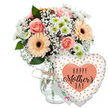 A warm and celebratory Mother's Day scene is captured here, as if a freshly delivered gift has just been set down on a bright North Watford kitchen counter. The background is a clean, uninterrupted white, which makes every colour in the flowers and balloon appear vivid and distinct, like a spotlight on a special moment. At the centre of the composition stands a clear glass vase with gentle curves and a faintly textured surface, half-filled with water that slightly magnifies the stems inside. Around the neck of the vase, a slim band of rustic twine is tied in an understated bow, lending an artisan, homely feel familiar to those who enjoy local deliveries around St Albans Road and the residential streets nearby. Rising from the vase is a lavish, rounded bouquet of fresh-cut blooms in soft, welcoming shades. Several peach-pink roses, open but not yet fading, display layers of velvety petals spiralling from tight centres. Interspersed among them are cheerful peach gerbera daisies whose wide, smooth petals surround dark brown to black centres, providing a striking contrast that draws the eye. Filling the spaces between are airy clusters of white baby's breath, their tiny flowers creating a gauzy halo, while small white or very pale green chrysanthemums contribute compact, button-like blooms that add substance and gentle brightness. Lush greenery in deep tones frames the pastels, giving depth and structure so the arrangement appears both full and naturally balanced. To the right of the bouquet, attached discreetly, a heart-shaped Mylar balloon seems to float. Its surface is crisp white, scattered with metallic gold and silver polka dots that shimmer softly. In the middle, a peach-toned heart shape encloses the words Happy Mother's Day written in elegant black script, the flowing lines echoing the curves of the rose petals. Together, the flowers, balloon, and a barely visible white card tucked behind suggest a complete, thoughtful Mother's Day gift, perfect for surprising a mum anywhere in North Watford.